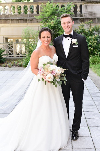 A bride wearing a long white gown and veil and a groom in a black tuxedo stand together outdoors