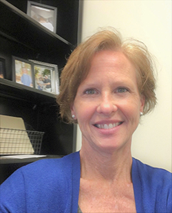 A white woman with short strawberry blonde hair smiles at the camera as she sits in front of a bookshelf