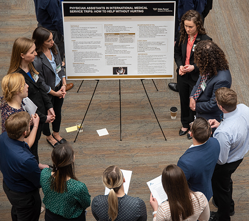 A group of young people stand in a circle around a poster listening to a single woman talk