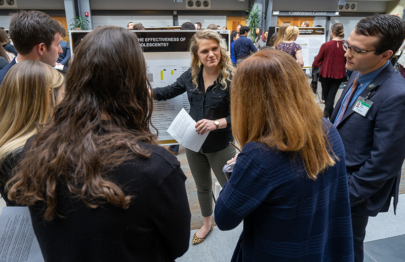 A young blonde woman gestures toward a research poster as she talks to a group of people