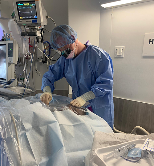 A person in blue scrubs and PPE works on a draped object, presumably a patient in a hospital room