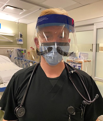 A white man with strawberry blond hair wearing a navy scrub shirt, mask and face shield, stands in a hospital