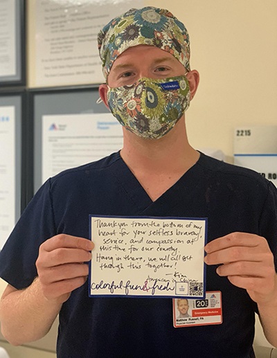 A white man in matching patterned mask and head covering, wearing a navy scrub shirt, holds up a handwritten postcard