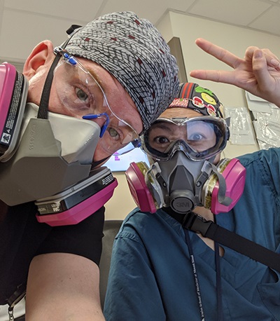 A man and woman wearing scrubs, head coverings, face shields and gray-and-pink respirators pose for the camera