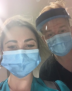 A man and woman, wearing scrubs and face masks and, for the man, a face shield, look into the camera for a selfie