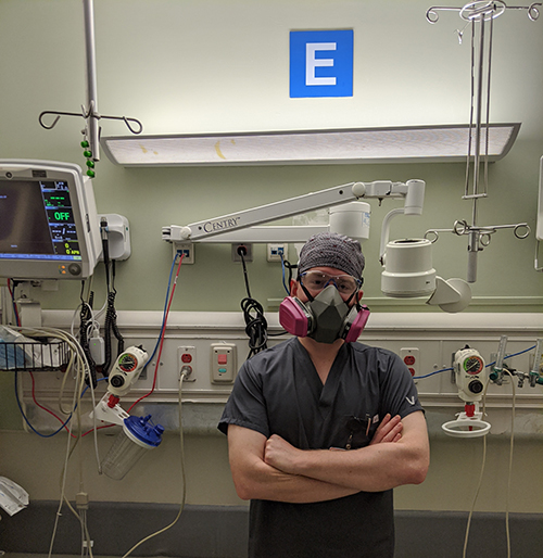 A white man wearing navy scrubs, head covering, respirator and face shield, stands in front of hospital equipment with arms crossed