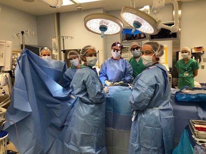A group of people in blue scrubs, head coverings, masks and gowns turn from the operating table to look at the camera