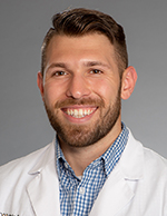 A white man with brown hair, beard and mustache wearing a white coat smiles for a photo