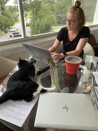 A woman with hair in ponytail and wearing glasses works on a laptop on a glass-topped table covered with notepads, drinks and a black-and-white cat