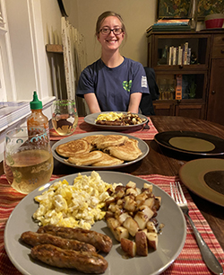 A smiling woman sits at the far end of a table loaded with plates filled with breakfast food