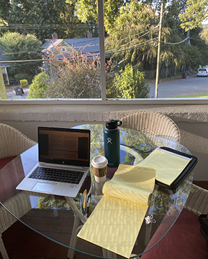 A laptop, yellow notebook, coffee and water bottle sit on a glass-topped table on a screened porch