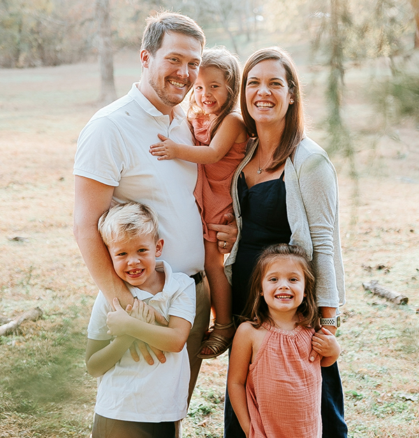 A man, a woman and three young children stand outdoors with arms around each other and smile at the camera
