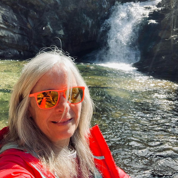 Woman with long white-gray hair wearing red jacket and red sunglasses takes selfie in front of a waterfall while kayaking on a river
