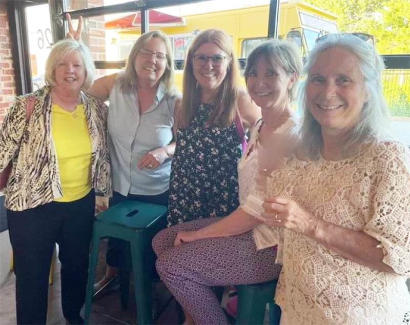 Five older white women sit and stand together and smile at the camera