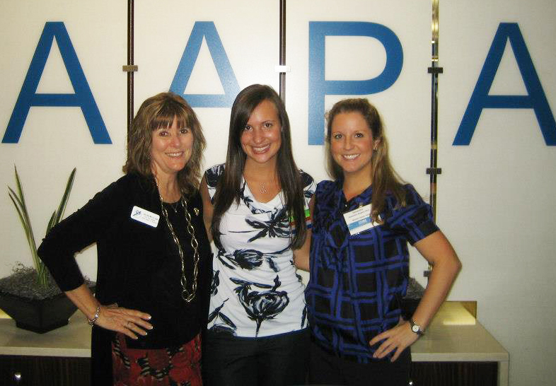 Three women stand in front of a sign reading 'AAPA' and smile for the camera
