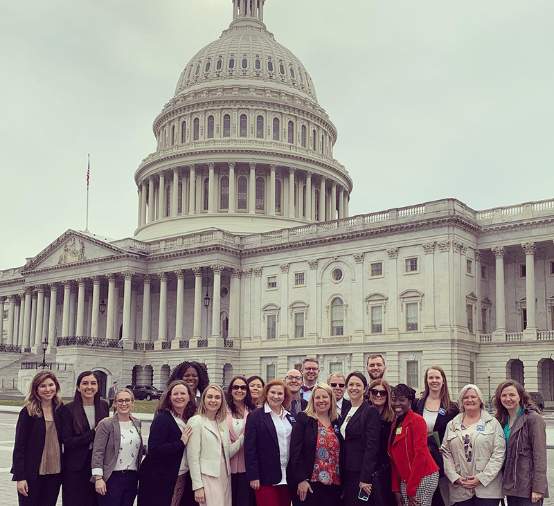 A large group of men and women stand in front of the Capitol Building in Washington DC and pose for a group photo under an overcast sky