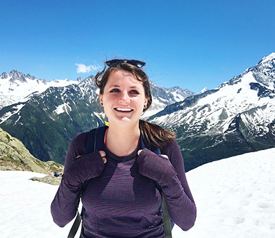 Woman with long dark hair in a ponytail and wearing a long-sleeve purple shirt and backpack stands in a snowfield with a snow-capped mountain range behind her