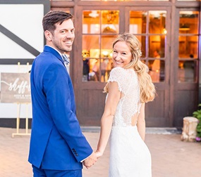 A man in a blue suit and a woman in a wedding dress hold hands and look back over their shoulders at the camera in front of a door