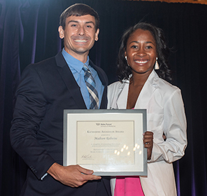 A man wearing a dark blue suit and a younger Black woman and wearing a white coat smile as they hold a framed award.