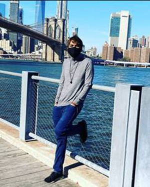 A dark-haired man leans back against a fence with a river and the New York City skyline behind him. He is wearing jeans, a gray shirt and a black face mask