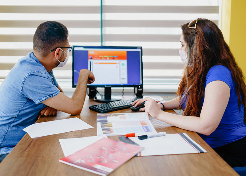 Hands of two men, one holding a pen and pointing at writing on a piece of paper on a table, lying between two laptop computers