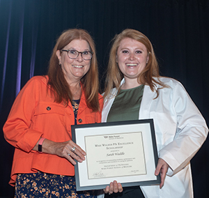 A woman in an orange jacket and a younger woman and wearing a white coat smile as they hold a framed award.