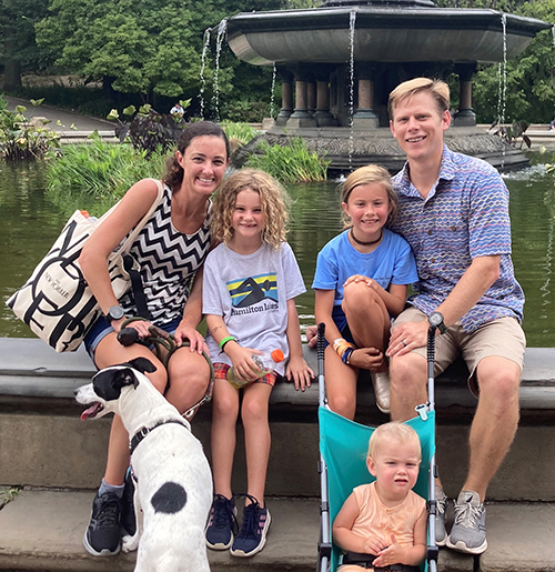 A family group of a mom, dad, three young children and a black-and-white dog sit on the edge of a large fountain and smile at the camera