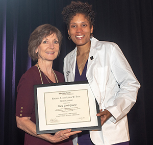 Two women, one older and white, the other Black and wearing a white coat, smile as they hold a framed award.
