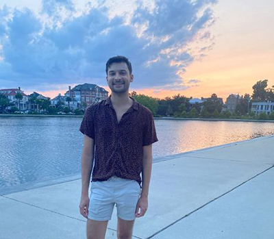 Man with dark hair wearing a buttondown short-sleeve shirt and shorts stands on a waterfront with houses and a sunset behind him