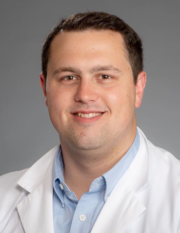 A man with dark hair wearing a lab coat and smiling at the camera.