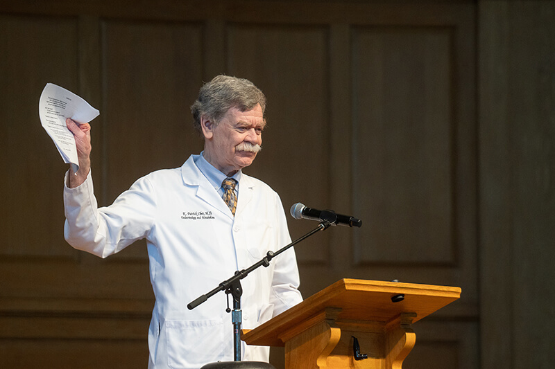 An older man standing at a podium speaking holding up a piece of paper.