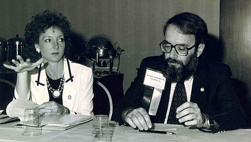 A black and white photo of a woman speaking at a summit while sitting next to a man.