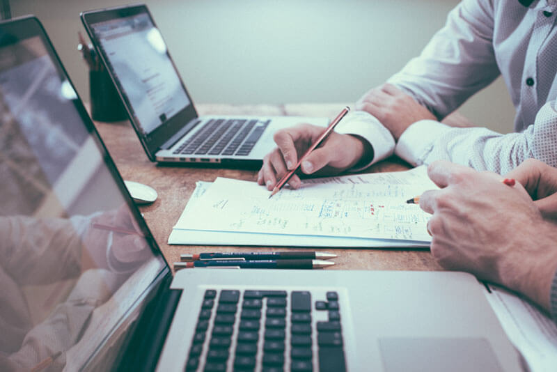 Two gray laptops on a desk with a stack of paper notes in-between them being written on.