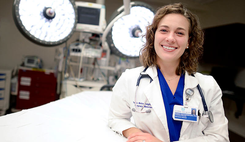 Hannah Cox Muthersbaugh in blue scrubs and a lab coat smiling in front of a surgical table and lights.