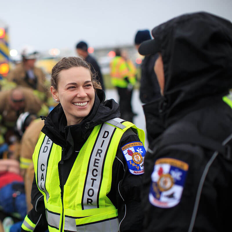 Hannah Cox Muthersbaugh a black uniform and neon green reflective vest speaking with a person in a hooded black uniform.