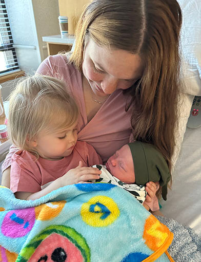 A baby in a pink shirt holding a sky-blue blanket being held by a brown-haired woman in a pink shirt.