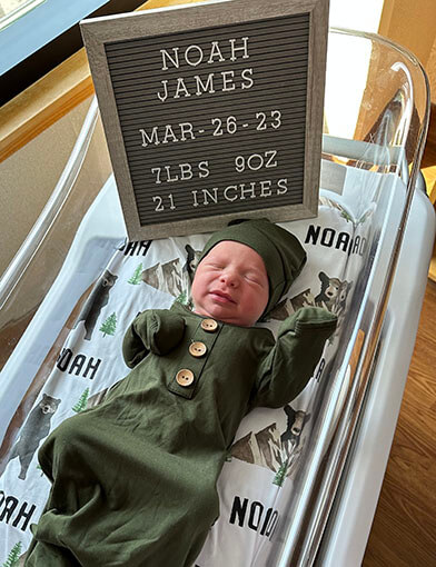 A baby in a green had and outfit lying down in front of a sign saying "Noah James Bates".