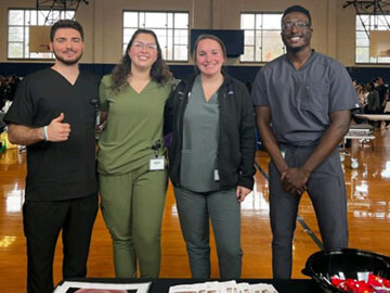 Two men and women in scrubs smiling in a group behind a table with paper handouts on it.