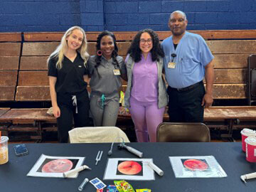One man and three women in scrubs smiling in a group behind a table with paper handouts on it.