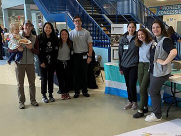 A group of medical students standing for a group picture near a staircase and a table with a display of medical information on it.