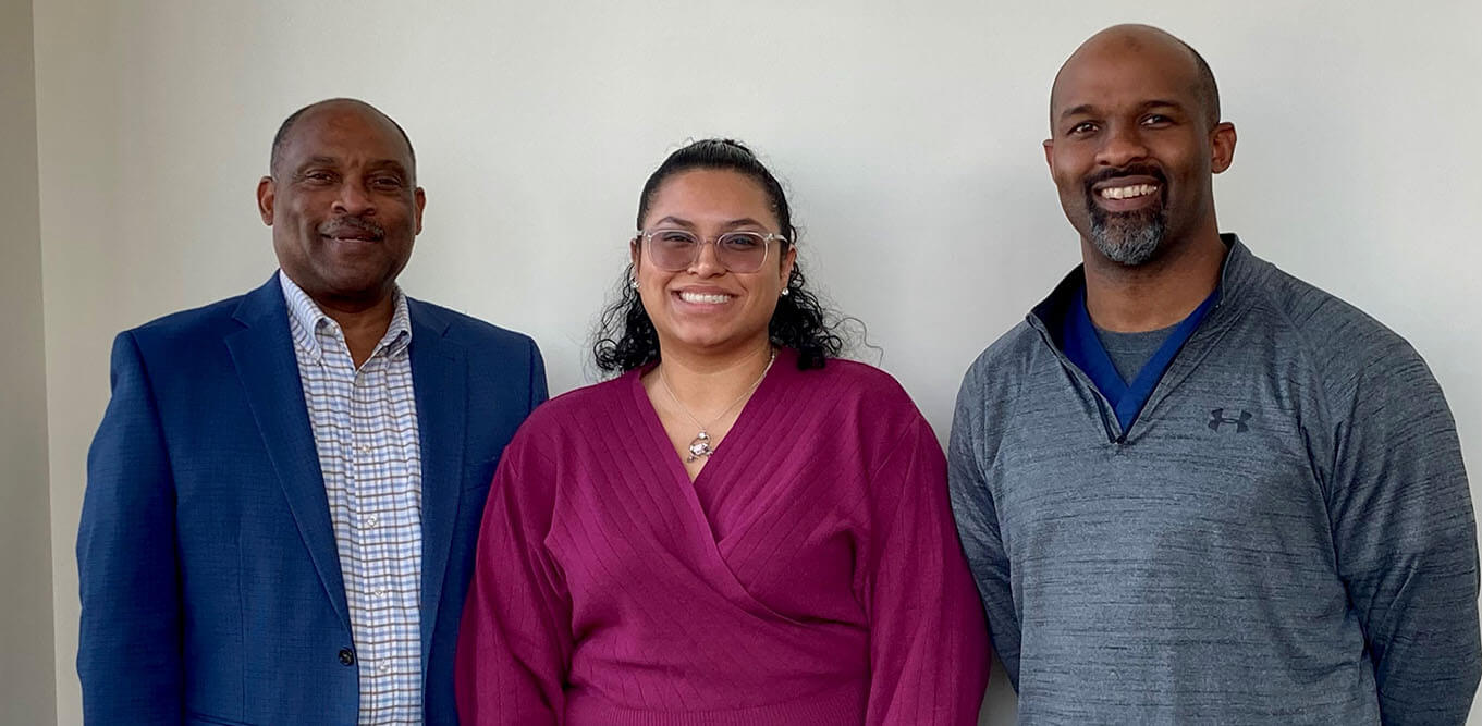 Robert Wooten and Aaron Wooten, who are wearing a blue suit and a gray long sleeve shirt pose for a photo with Jesseca Garcia.