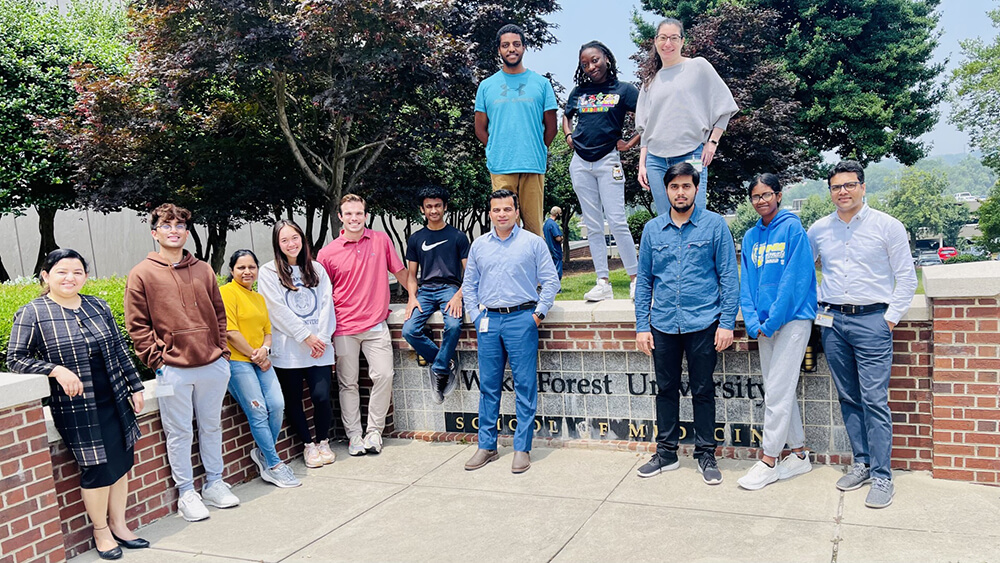 A group of men and women in casual attire smile and pose in a group photo on and near an outdoor brick and stone sign.