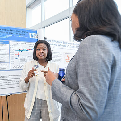 Two women talk next to a large board with research findings, charts and graphs .