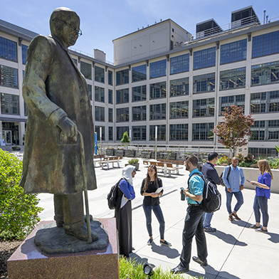 Courtyard of large, light-colored building with windows and multiple people standing and talking while holding books with the Bowman Gray statue in the foreground 