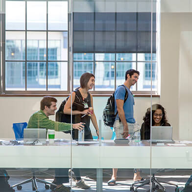 Two men in green and blue t-shirts and two women in a black dress and black shirt gathering in a conference room and talking.