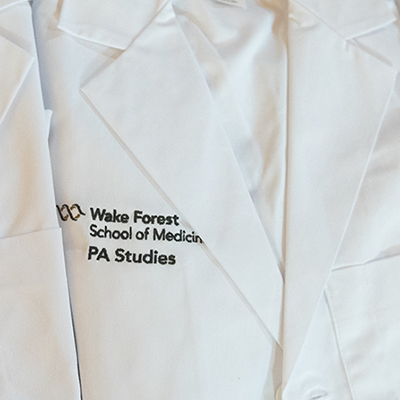 A row of neatly folded and pressed white coats embroidered with 'Wake Forest School of Medicine PA Studies' sit on a black-clothed table