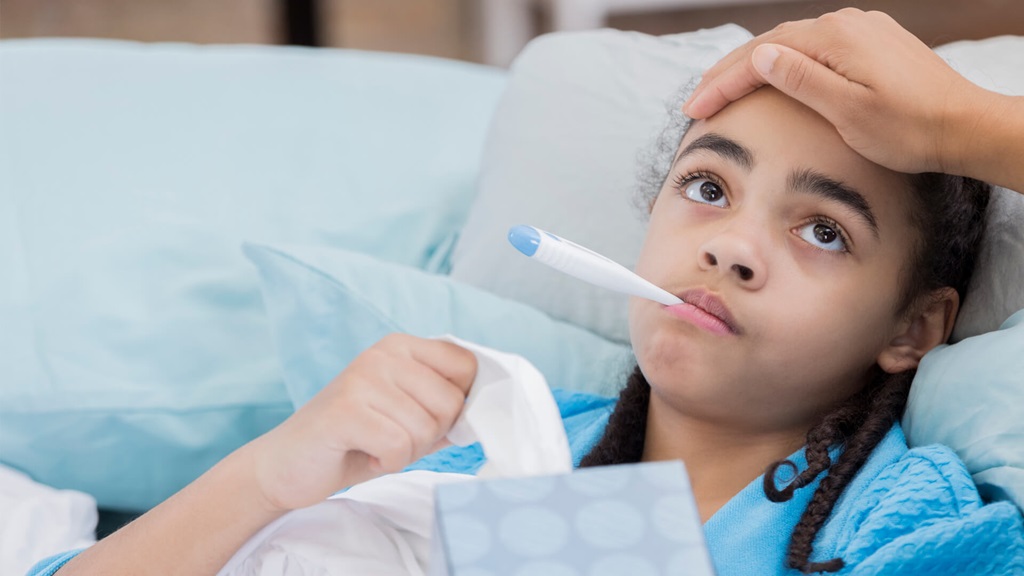A sick child resting in bed while an adult places a hand on their forehead.