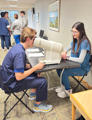 A young man sitting in a chair taking a vision test administered by a young woman in blue scrubs.