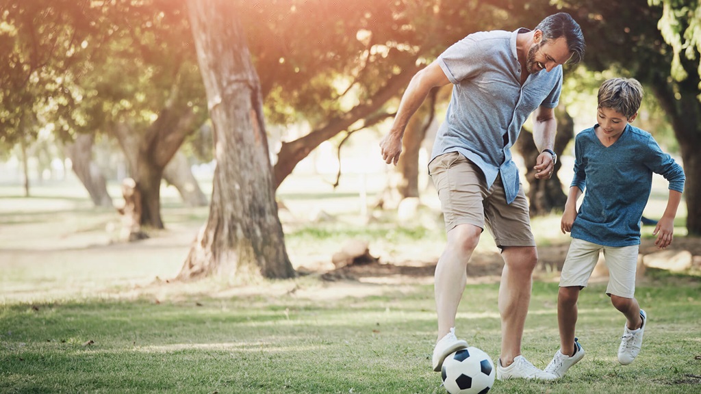 A young boy in a blue shirt and tan shorts playing soccer with his father.