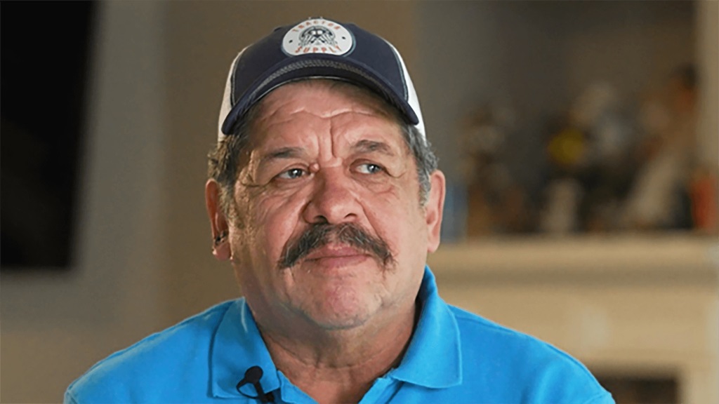 A man wearing a blue shit and a hat sitting in front of the camera.